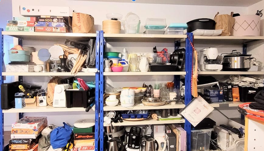 Fotografiet visar tre lagerhyllor som är fyllda med massor av föremål. Till exempel skålar, sällskapsspel, grytor, symaskin, vattenkokare med mera.